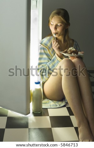 Young womanl eating cake beside fridge. She\'s looking at somethings in fridge. Front view.