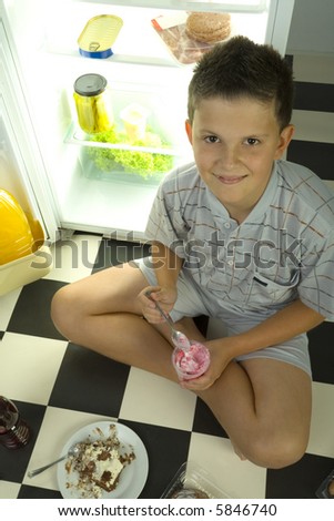 Young boy sitting by the fridge and eating ice cream. Looking at camera. High angle view