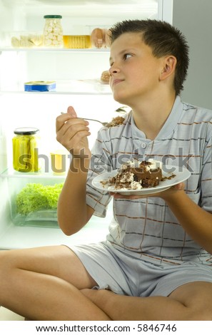 Young boy sitting beside fridge and eating cake. Front view