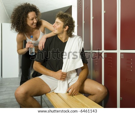 Young couple, sitting on bench and talking in gym's locker room. Man is holding towel, woman holding bottle of water. They're looking at each other. Front view