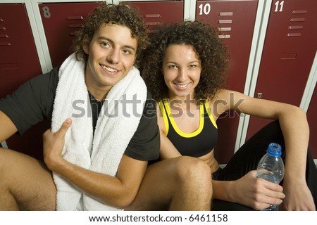 Young couple, sitting in gym\'s locker room in front of lockers. Man is holding towel, woman holding bottle of water. They\'re looking at camera. Front view