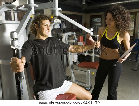 Young couple, working out in gym. Man is sitting and picking up dumbbell. He looks tired. Woman is standing close by man. Smiling and looking at him