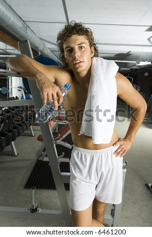 Young tired man standing in gym with bottle of water and towel. Looking at camera, front view