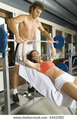 Young couple, working out in gym. Man is helping woman. Woman is lying on bench and picking up dumbbell. Smiling and looking at man. Side view