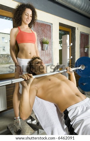 Young couple, working out in gym. Man is lying on bench and picking up dumbbell. Woman is standing behind him. Side view