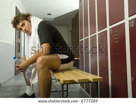 Young, tired man sitting on bench in locker room. Holding bottle of water in hand and towel on neck. Side view. Looking at camera