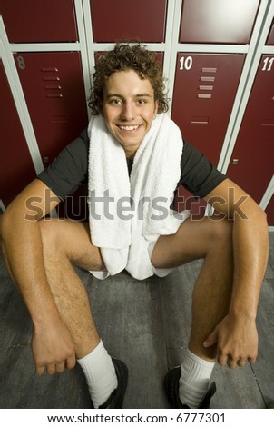 Young, tired but happy man sitting on the floor in locker room. Holding towel on neck. Front view. Looking at camera