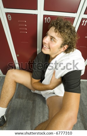 Young, tired but happy man sitting on the floor in locker room. Holding towel. High angle view. Looking at something