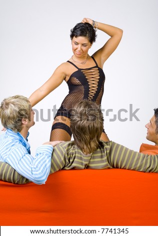 Three young men sitting back on orange couch in front of stripteaser. They are looking at her. She is touching one of them