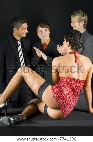 Three businessmen standing in front of woman in dark room. They are smiling and looking at woman. Woman is sitting on table. Front view