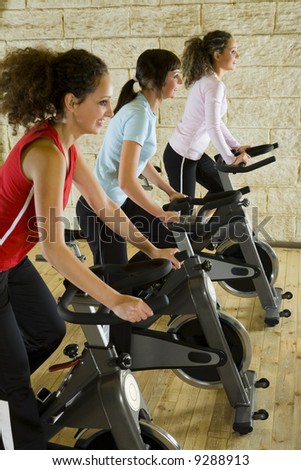 Three happy, young women working out on exercise bicycle at the gym. Focus on woman in red shirt. Side view.