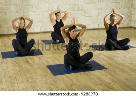 A group of women making exercise with uprised hands over heads and sitting with crossed legs on yoga mat. They\'re smiling and looking somewhere. Focus on the woman in pink shirt. Front view.