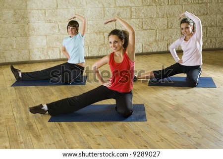 A group of women making stretching exercise on yoga mat. They\'re smiling and looking somewhere. Focus on the woman in red shirt. Front view.