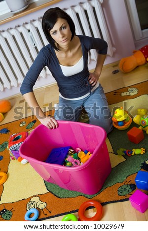Young woman kneeling at container with toys and assembles strewn toys. High angle view.