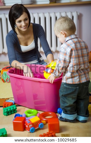 Young woman with baby boy during plaing. Woman showing toy to baby. They are at container with toys. Baby standing back to camera. Front view.