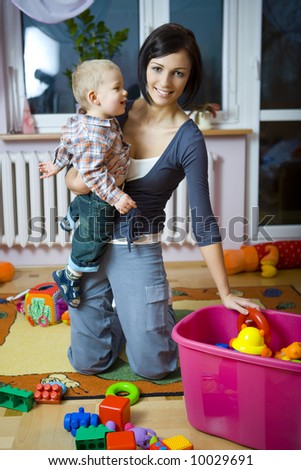 Young woman with baby boy during plaing. Woman holding baby on hand taking toy from container. Front view.