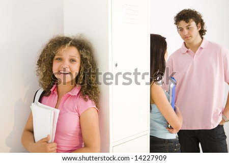 Theree schoolmate standing by school locker. Girls leaning for shool locker and holding notebooks. Boy talking with girl in blue shirt. Focuset on smiling girl in pink shirt.