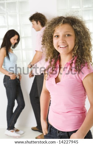Three friends standing near window. A boy talking with girl in blue shirt. A girl in pink shirt smiling and looking at camera. Focused on girl in pink shirt.