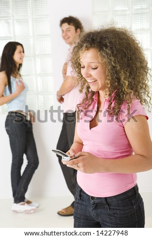 Three happy friends standing near window. A boy talking with girl in blue shirt. A girl in pink shirt holding mobile phone. Focused on girl in pink shirt.