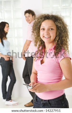 Three happy friends standing near window. A boy talking with girl in blue shirt. A girl in pink shirt holding mobile phone and looking at camera. Focused on girl in pink shirt.