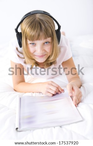 Young smiling girl lying on a bed with earphones. She's browsing a journal.