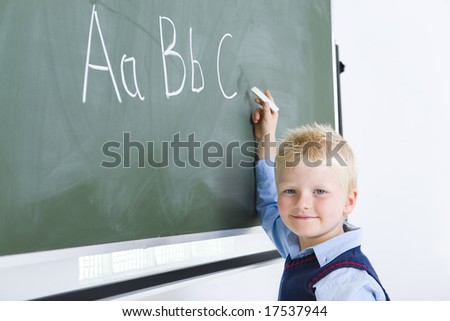 Young smiling boy writing alphabet on chalkboard. He\'s looking at camera.