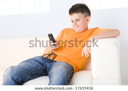 Young boy sitting on couch and watching something in cellphone. Front view.