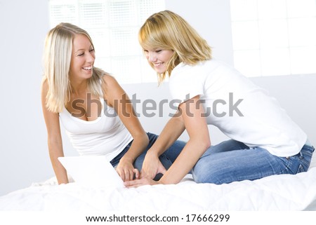 Two girl friends sitting on bed and do something on the laptop. Front view.