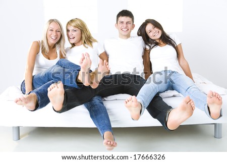 Group of young smiling friends sitting on bed with extended legs. They\'re looking at camera. Front view.