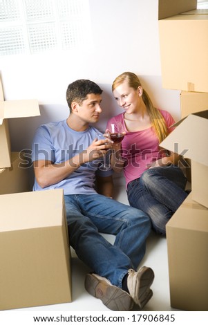 Young couple sitting between cardboard boxes and drinking wine. They're looking at each other's. Front view.