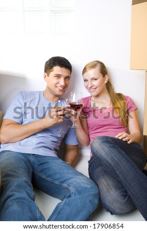Young couple sitting between cardboard boxes and drinking wine. They\'re smiling and looking at camera. Front view.