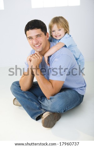 A man sitting on the floor with cross-legged and his daughter hugging him. They're smiling and looking at camera.