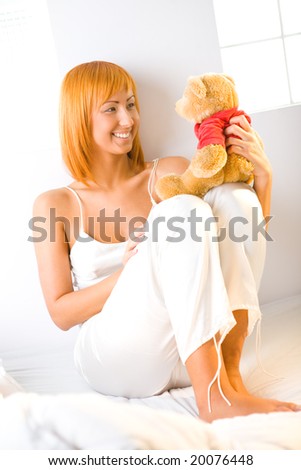 Young red-haired woman dressed pyjamas sitting on bed and holding teddy bear. She\'s leaning against a wall. She\'s smiling and looking at teddy bear.