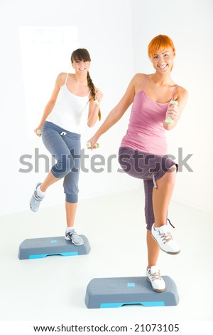 Two young women doing exercise on aerobic step. They\'re looking at camera. Front view.