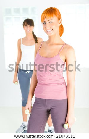 Two young women doing fitness exercise with dumbbells. They\'re looking at camera.
