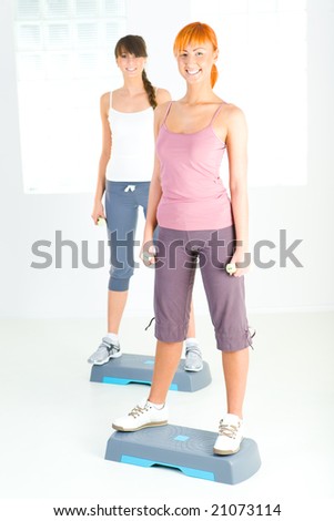 Two young women doing exercise on aerobic step. They\'re looking at camera. Front view.