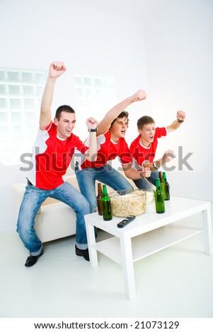 Three happy sport\'s fans get up from couch with raised hands.