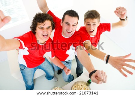 Three happy sport\'s fans get up from couch with raised hands. They looking at camera. High angle view.