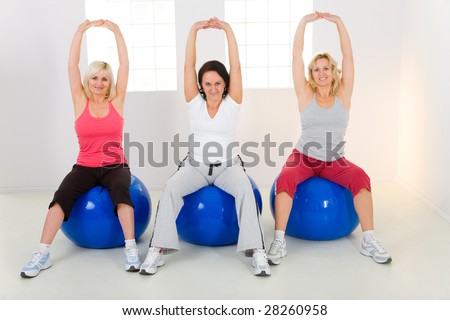 Women dresset sportswear working out on fitness ball. They have raised hands. They\'re smiling and looking at camera. Front view.