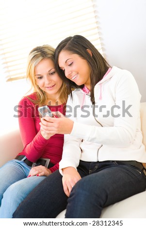Two girls looking at cellphone and smiling. They\'re sitting on couch.