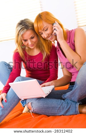 Two young women sitting on bed and doing something on laptop. One of them talking by cellphone.
