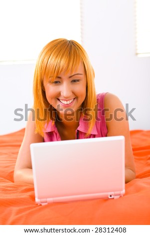 Young woman lying on bed and doing something on laptop. Front view.