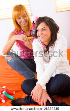 Young woman curling hair her friend. They\'re sitting on bed. Front view.