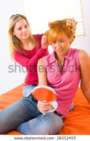 Young woman styling hair her friend. Red-haired woman with curlers on head holding hand glass.