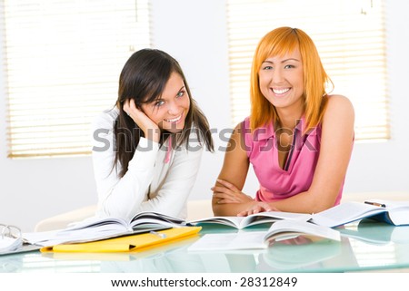Two girls sitting at the table and doing homework.They're smiling and looking at camera. Front view.