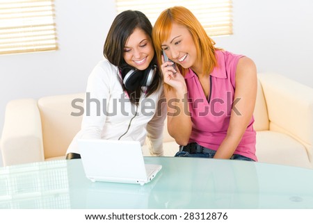 Two girls sitting on couch at table. One of them speaking by cellphone. They're smiling and looking on laptop. Front view.