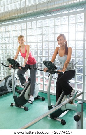 Young women working out  on bike at gym. They're smiling and looking at camera.