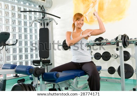 Happy woman sitting on exercising bench. She's smiling and pointing at her biceps.