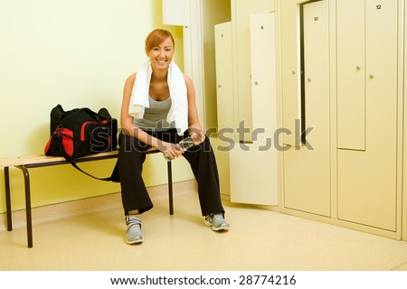 Young woman with towel sitting on bench in locker room. She\'s holding bottle of water.