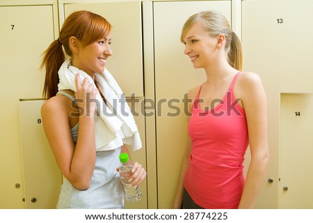 Two women stand in locker room and talking. One of them holding towel and bottle of water.